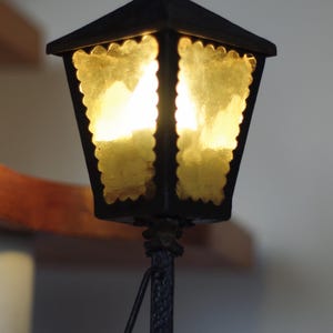 May include: A black metal lantern lamp with a square base and a textured post. The lantern features four panels of amber-colored, scalloped-edge glass that emit a warm glow. A small finial sits atop the roof, and a white power switch is on the base.