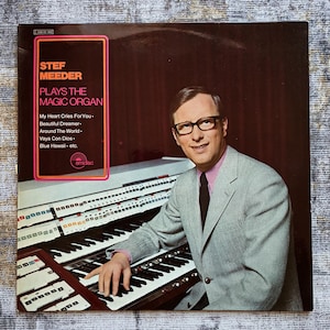 Könnte beinhalten: Albumcover von "Stef Meeder spielt die Magic Organ". Das Bild zeigt einen Mann im grauen Anzug und Brille an einer Orgel. Der Albumtitel und die Titelliste sind in einem rosa-braunen Rahmen dargestellt. Das emidisc-Logo ist sichtbar.