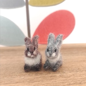 Puede incluir: Dos pequeños conejos de fieltro hechos a mano. Uno es marrón y blanco, el otro gris y blanco. Están sobre una superficie de madera, con un diseño de hojas decorativas en el fondo. Los conejos miden aproximadamente 5 cm de altura.
