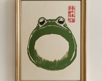 May include: A framed illustration of a green frog in a circle shape, with its eyes and mouth visible. A red Japanese character stamp is in the top right corner.