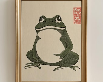 May include: A framed print of a green frog with a white belly, sitting on a beige background. The print has a red stamp in the top right corner with Japanese characters.
