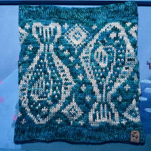 May include: A knitted cowl in shades of teal, turquoise, and white, featuring a geometric pattern. The design includes stylized floral or tree-like motifs. A small tag with paw prints and initials is visible on the lower right corner. The cowl is likely handmade.