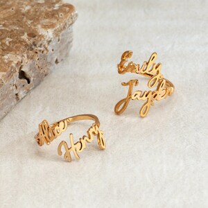 May include: Two gold-coloured metal rings featuring custom script lettering. One ring displays the names Emily and Jayden, while the other ring features the names Alice and Henry. The rings are crafted with a polished finish and open bands.