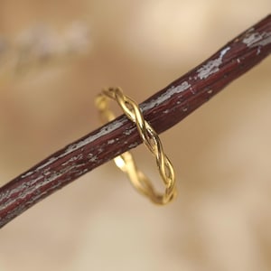 May include: A thin, polished gold ring featuring a delicate twisted rope design. The band is constructed from two intertwined strands of metal, creating a continuous spiral pattern that encircles the entire circumference of the circular ring.