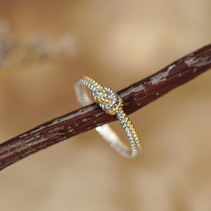 May include: A silver and gold-toned knot ring. The ring is set against a blurred background and a brown branch. The ring's knot design is in the center, with a gold-toned knot and silver-toned band.