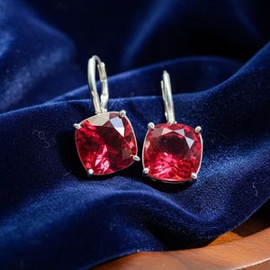 Op de afbeelding: Een paar zilverkleurige oorhangers met grote, vierkant geslepen, robijnrode edelstenen. De oorbellen zijn geplaatst tegen een diepblauwe fluwelen achtergrond, wat een opvallend contrast creëert.