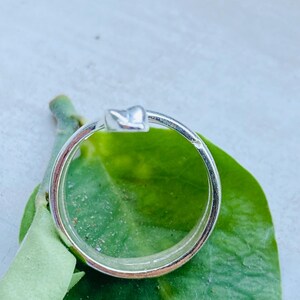 May include: A silver ring with a heart-shaped design. The ring is set against a green leaf, with the ring's band and heart detail clearly visible. The ring is a simple, elegant piece of jewelry.
