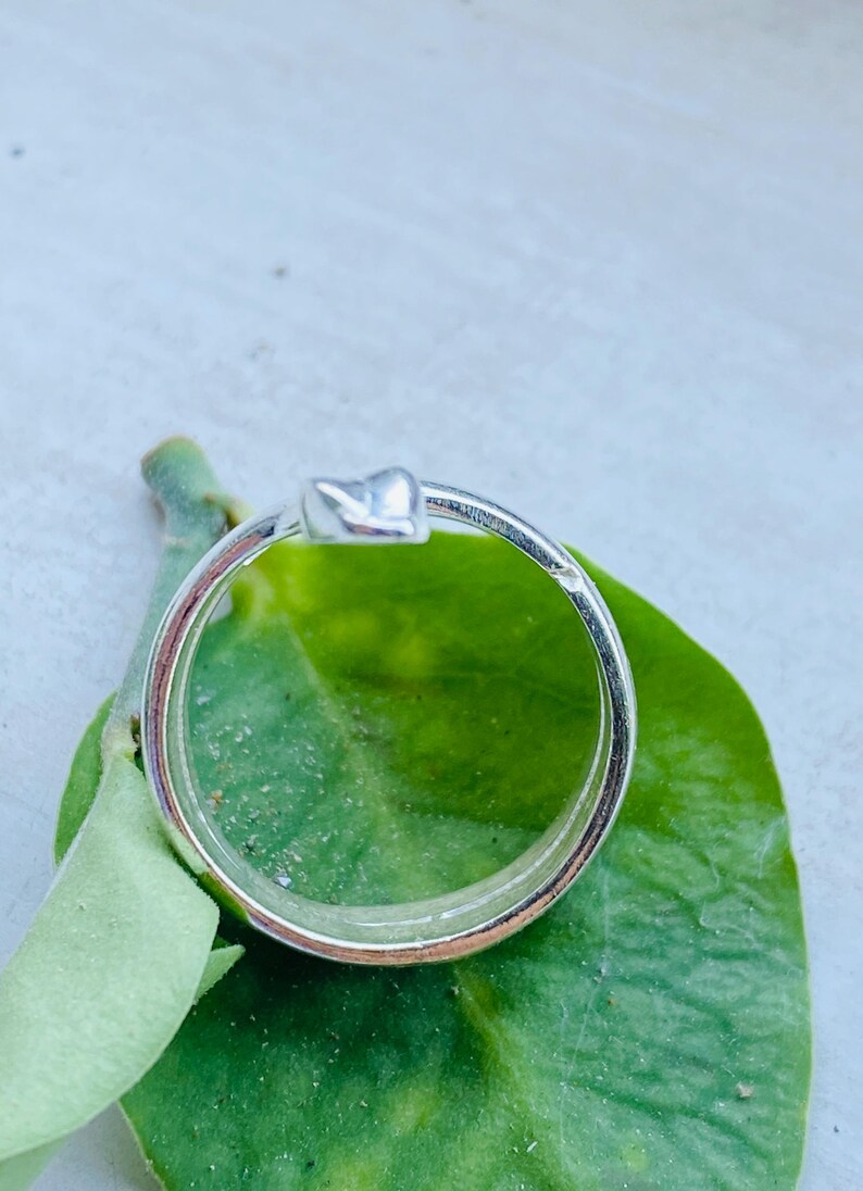 May include: A silver ring with a heart-shaped design. The ring is set against a green leaf, with the ring's band and heart detail clearly visible. The ring is a simple, elegant piece of jewelry.