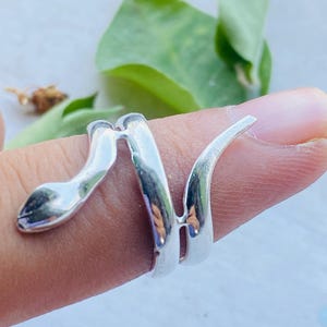 May include: A silver-colored snake ring wraps around a finger. The ring's design features a snake head and a spiraling body. The ring is made of a shiny, reflective metal, and the snake's head is detailed with a slight curve.