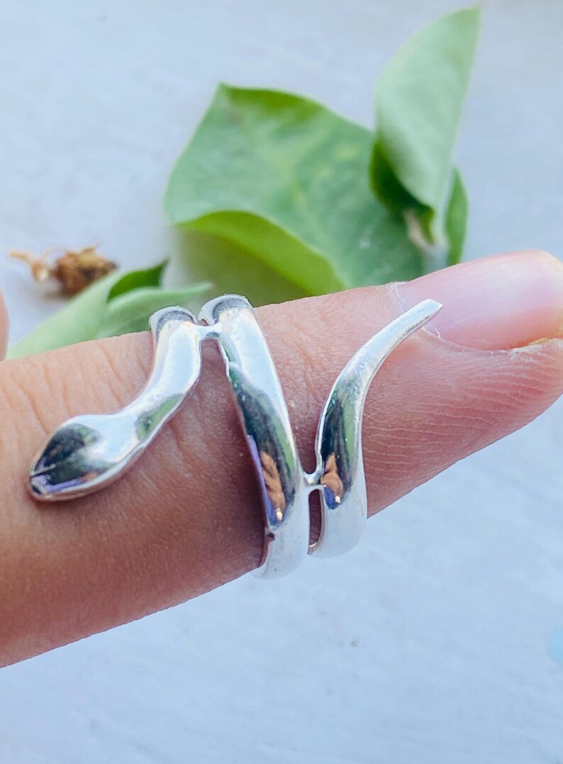 May include: A silver-colored snake ring wraps around a finger. The ring's design features a snake head and a spiraling body. The ring is made of a shiny, reflective metal, and the snake's head is detailed with a slight curve.