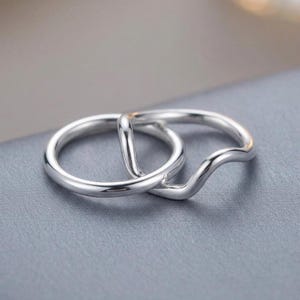 May include: Two interlocking silver rings with a polished, reflective finish. One ring features a smooth, circular band, while the other ring has a distinct wave-like curve along its circumference, creating a layered and connected appearance.
