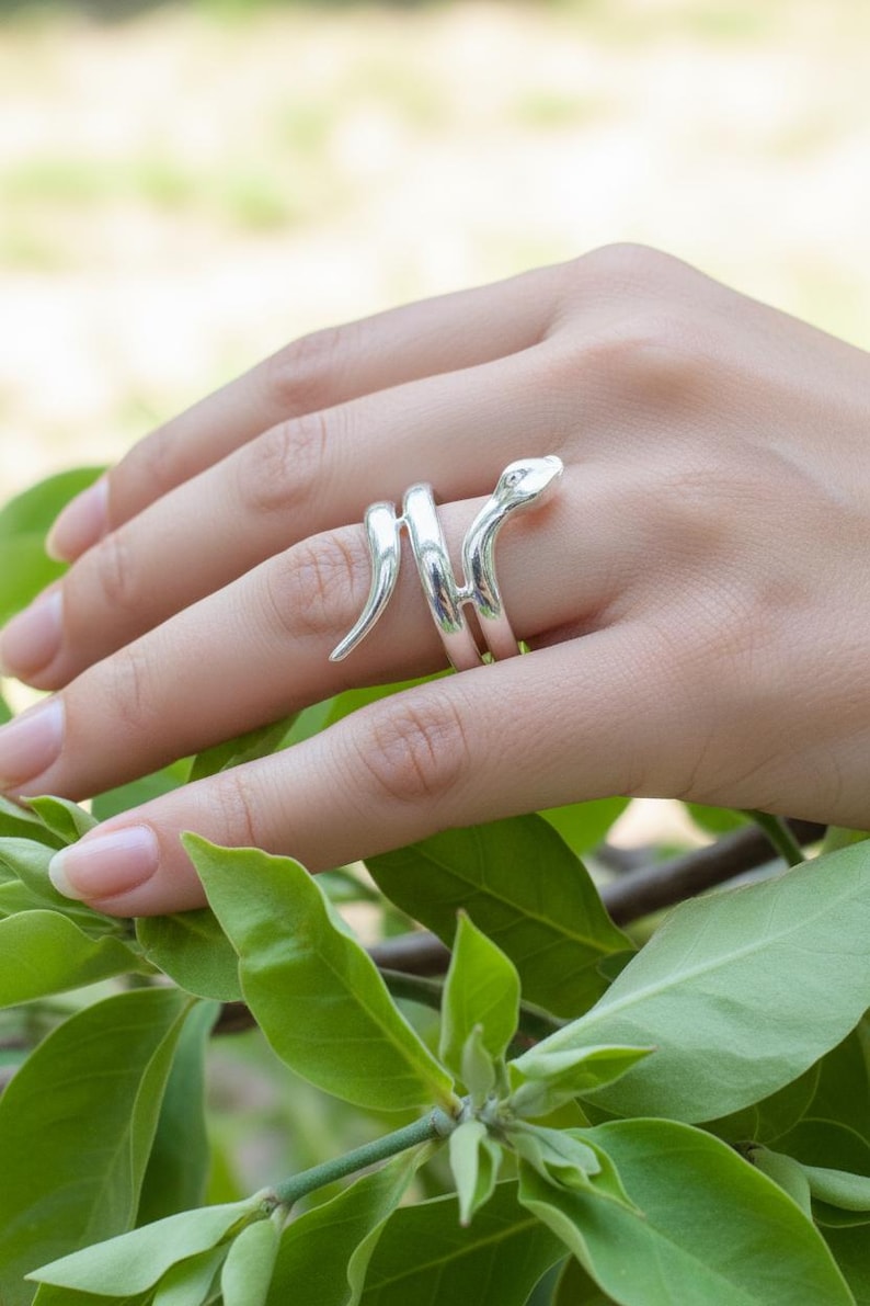 May include: A silver snake ring coiled around a finger. The ring features a detailed snake head and tail, with the body of the snake wrapping around the finger. The ring is set against a background of green leaves.