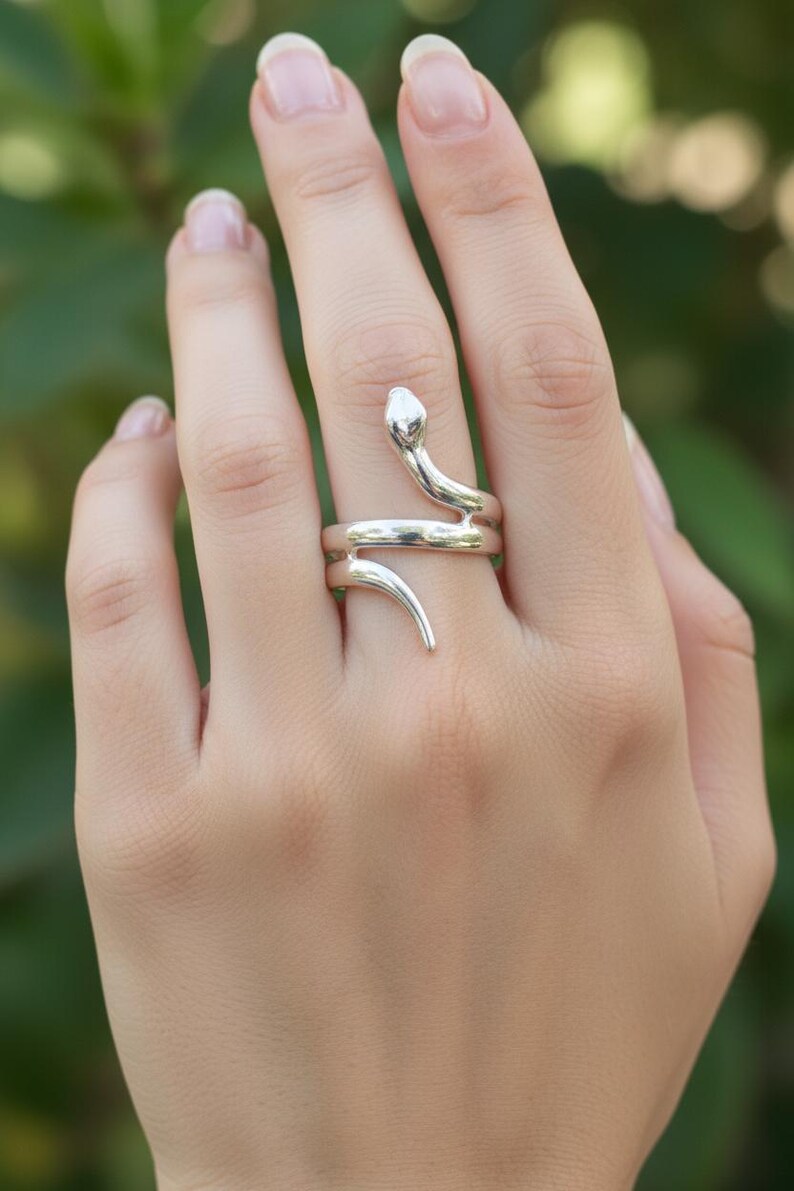 May include: A silver-colored ring in the shape of a snake, with the snake's head and tail curving around the finger. The ring is worn on a hand, with a blurred green background.