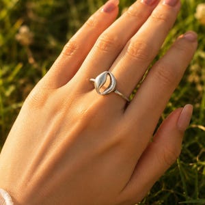 Può includere: Un anello color argento caratterizzato da un piccolo ciondolo a forma di conchiglia di ciprea liscia, centrato su una sottile fascia metallica lucida. La conchiglia presenta una distinta apertura longitudinale con dettagli finemente rigati.