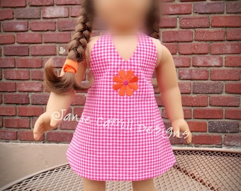 May include: A doll wearing a pink and white gingham dress with an orange flower embroidered on the front. The doll has brown hair and is wearing white sandals.