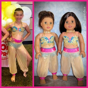 May include: Two dolls wearing matching turquoise, gold, and pink costumes. The costumes feature a jeweled top, a pink sash, and sheer gold pants. The dolls have brown hair and are standing on a light wood floor.