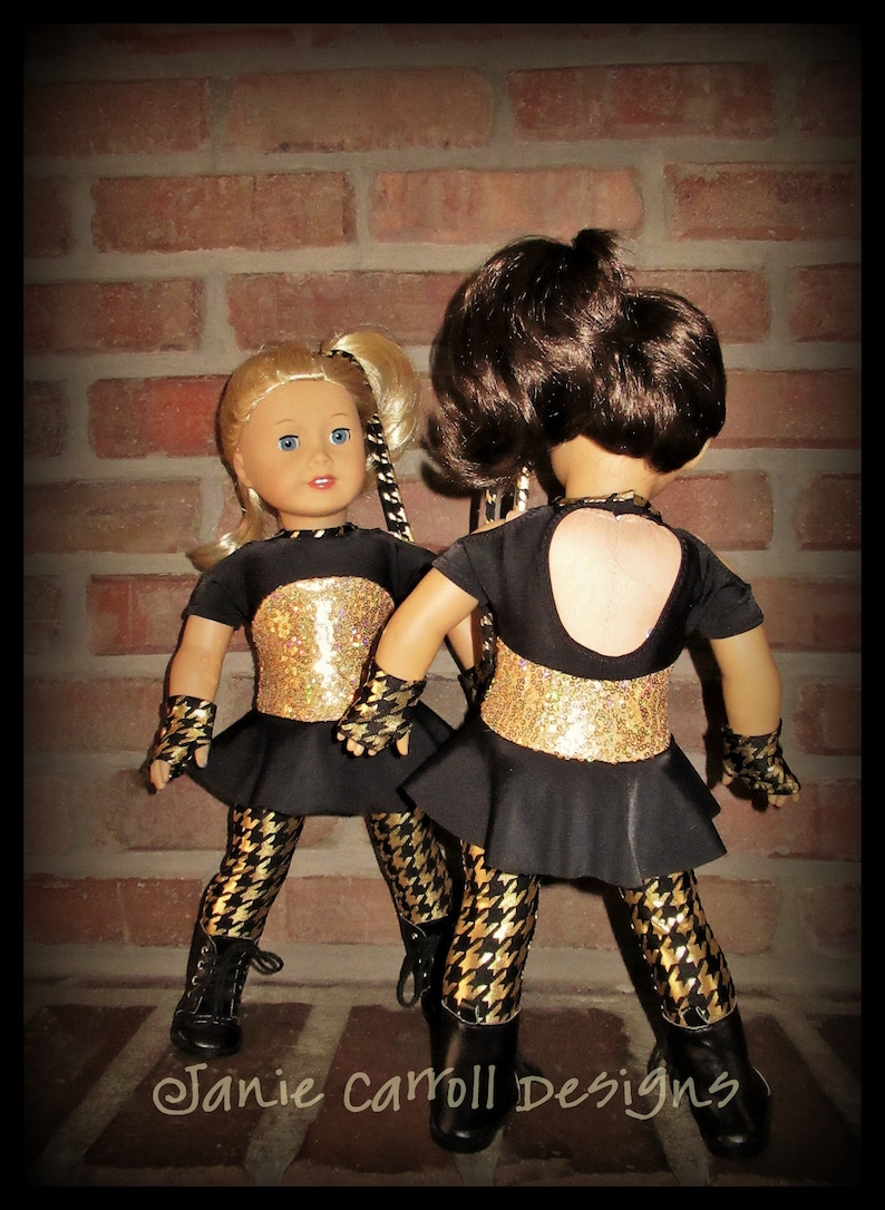May include: Two dolls wearing black and gold dance costumes with black and white patterned leggings. The dolls are standing in front of a brick wall. The text "Janie carroll Designs" is visible at the bottom of the image.