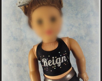 May include: A black cheerleading outfit with silver glitter accents. The top has a scoop neck and the word "Reign" in silver glitter with rhinestones. The shorts have a silver glitter waistband. The outfit is worn by a doll with brown hair and a silver glitter bow.