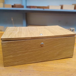 Könnte beinhalten: Eine Holzkiste mit einem Scharnierdeckel und zwei Metallverschlüssen. Die Kiste ist aus hellbraunem Holz mit natürlicher Maserung.