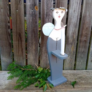May include: A wooden angel candle holder painted in white and gray, with a red beaded necklace and a silver halo. The angel holds a white candle. The holder is standing on a wooden surface in front of a wooden fence.