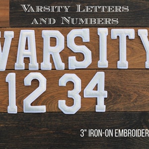 May include: White iron-on embroidered varsity letters and numbers, 3 inches tall. The letters spell out "VARSITY" and the numbers are 1, 2, 3, and 4. The letters and numbers are on a wood background.