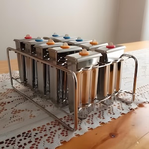 Może przedstawiać: Zestaw ośmiu form do lodów na patyku ze stali nierdzewnej z kolorowymi pokrywkami, ułożonych w metalowej kratce. Formy są przeznaczone do robienia domowych lodów na patyku.