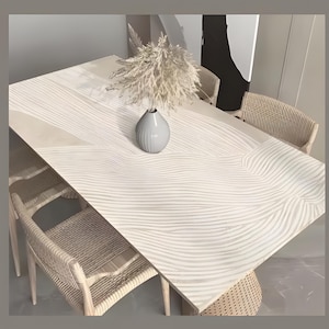 Może przedstawiać: Biały stół jadalny z falistą, fakturowaną powierzchnią. Stół jest otoczony czterema plecionymi krzesłami i wazą z suszoną trawą pampasową.