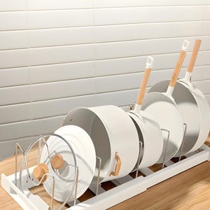 May include: White pots and pans with wooden handles are organized in a white metal rack. The rack is on a wooden surface.