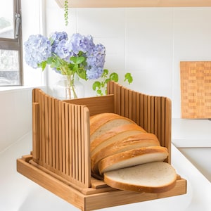 Könnte beinhalten: Ein Holz-Brotschneider mit einem herausnehmbaren Tablett und einem integrierten Brotmesser. Der Brotschneider ist aus natürlichem Bambus gefertigt und hat eine hellbraune Farbe. Ein Laib Weißbrot ist geschnitten und auf dem Tablett platziert.