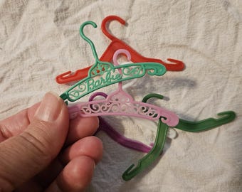 May include: A collection of miniature plastic doll clothes hangers in red, green, light pink, dark green, and purple. The light green hanger features the word Barbie embossed in script lettering with decorative scrollwork details on the frame.