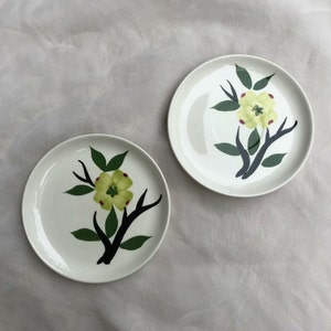 May include: Two white ceramic plates with a black branch and green leaves with a yellow flower design.