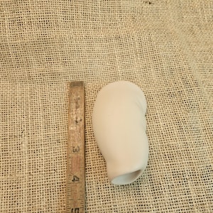 May include: A smooth, matte, off-white ceramic doll arm part with a rounded shoulder and a hollow opening at the wrist. A wooden ruler is placed alongside the object, showing it measures approximately 3.5 inches in total length.