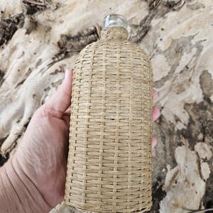 May include: A clear glass bottle encased in a woven, light brown wicker sleeve. The bottle has a clear glass neck and a woven texture. The bottle is held by a person's hand.