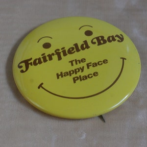 May include: Yellow smiley face button with brown text that reads "Fairfield Bay The Happy Face Place".