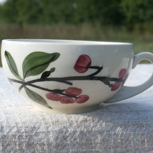 May include: Cream-colored ceramic teacup with a painted floral design. The cup features a black branch with green leaves and red berries. The handle is curved and the cup sits on a white surface.