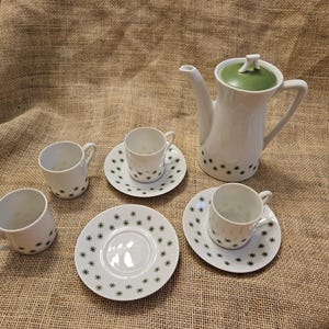 May include: White ceramic coffee set with a green-topped coffee pot, cups, and saucers. The set features a starburst pattern in black. The coffee pot has a curved spout and handle. The cups and saucers are small.