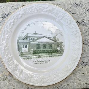 May include: White ceramic plate with a gold rim and a raised floral design. The center of the plate features a black and white illustration of a church building with the text "First Christian Church Sulphur Springs, Texas".