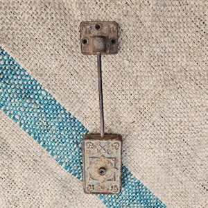 May include: A vintage, rusty metal door stop. The door stop has a rectangular base with an ornate design and a square top with mounting holes, connected by a thin rod. The base has the numbers "23" and "25" on it.