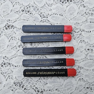 May include: A collection of five black Genuine Eversharp Leads cases with red caps. The text "GENUINE EVERSHARP LEADS" is printed in gold on each case. The cases are arranged on a white lace surface.