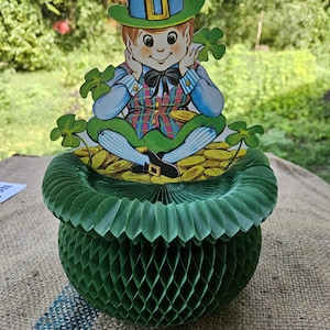 May include: A green honeycomb paper decoration featuring a leprechaun illustration. The leprechaun wears a green hat, a plaid vest, and a black bow tie, sitting atop a pile of gold coins surrounded by four-leaf clovers on a circular base.