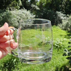 May include: A clear glass mug featuring a delicate etched design of pine needles and branches on the side. The mug has a sturdy handle and a smooth, transparent surface, showcasing a simple and functional design for holding beverages.
