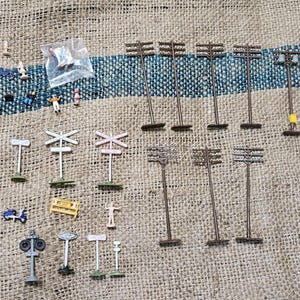 May include: A collection of miniature model train accessories including brown utility poles, white railroad crossing signs, various street lamps, and small painted plastic figures of people and vehicles arranged on a textured surface.