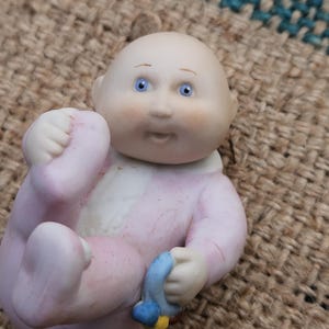 May include: A small porcelain figurine of a bald baby with blue eyes, wearing a light pink onesie with a white collar. The baby is in a reclining position, holding a small blue and yellow rattle in one hand and touching its foot with the other.