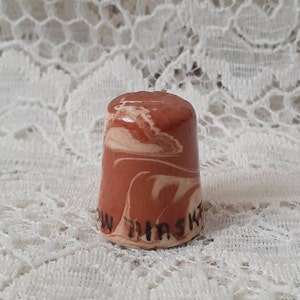 May include: A ceramic thimble with a marbled design in shades of brown and white. The thimble has the words "SNOW MASKA" printed in black. The thimble is a sewing accessory.