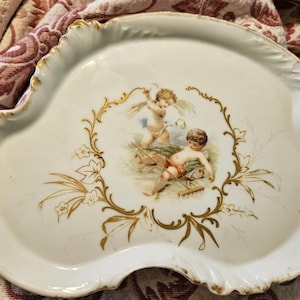 May include: White porcelain serving tray with a scalloped edge and gold trim. The center features a painted scene of two cherubs with a gold floral border. The tray has a unique, asymmetrical shape.