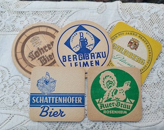 May include: Four different beer coasters with various designs. The coasters are round and square, with brown, yellow, and green backgrounds. The coasters feature blue and white text and logos, including "Johrer Bier", "Bergbrau Leimen", "Goldberg Pilsener", and "Auer-Brau Rosenheim".