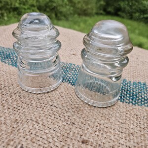 May include: Two clear glass insulators with a ridged design. The insulators have a rounded top and a cylindrical base. They are sitting on a burlap surface with a blue and white stripe.