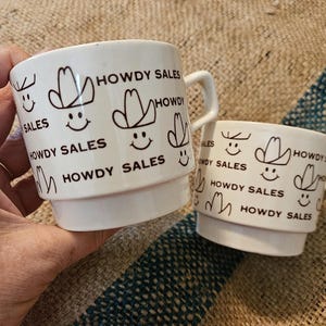 May include: Two white ceramic mugs featuring a repeating brown graphic print of a smiling face wearing a cowboy hat and the text HOWDY SALES. Each mug has a stepped base design and a side handle, with the text and graphics printed clearly.
