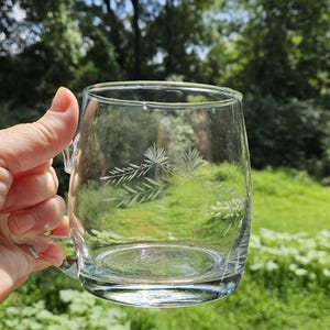 May include: A clear glass mug featuring a delicate etched design of pine needles and branches on the side. The mug has a smooth, rounded body with a sturdy handle attached to the side, showcasing a transparent finish and a simple, functional form.