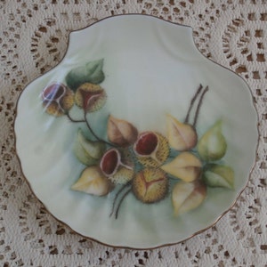 May include: A white porcelain shell-shaped dish with a gold rim. The dish is decorated with a hand-painted design of brown, yellow, and green leaves and chestnut-like fruits.